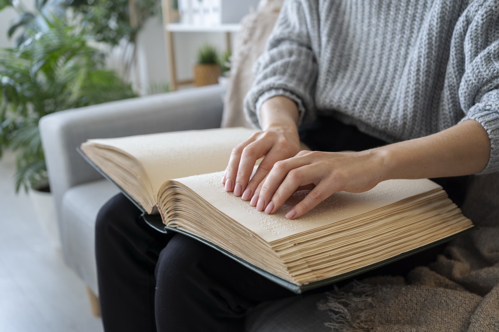 Auf dem Bild sieht man eine Person, die ein großes Buch in Brailleschrift liest. Die Person sitzt auf einem Sofa oder Sessel und trägt einen grauen, grob gestrickten Pullover und schwarze Hosen. Beide Hände sind auf den Seiten des Buches und tasten die erhabenen Braille-Punkte mit den Fingerspitzen ab. Die Seiten des Buches sind dick und cremefarben, was typisch für Braillebücher ist, da die erhabenen Punkte Platz benötigen. 
Im Hintergrund sieht man eine gemütlich eingerichtete Wohnumgebung mit grünen Zimmerpflanzen, einem hellen Raum und Möbeln in sanften Farben. Die Szene vermittelt eine ruhige und konzentrierte Atmosphäre..